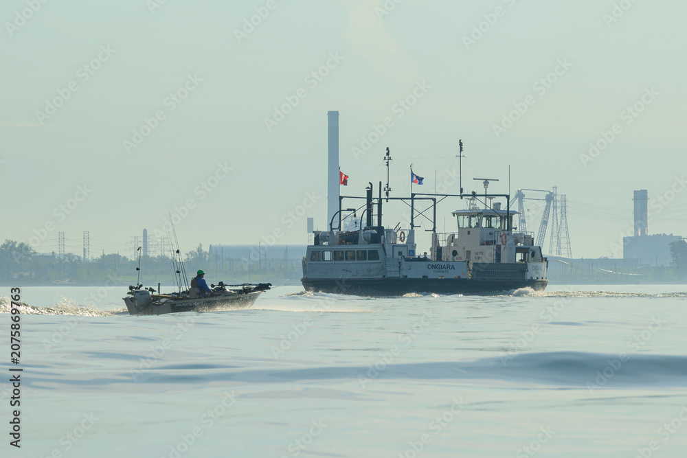 Fototapeta premium Ferry and small fishing boat crossing hazy harbour water near an industrial skyline at sunrise.