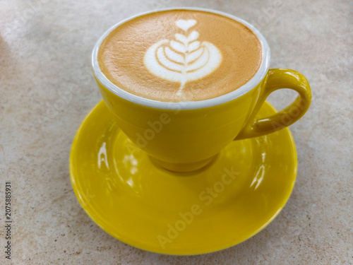 Coffee latte art with tulip pattern in yellow ceramic cup and saucer on textured table background. Hot cappuccino espresso beverage closeup view.