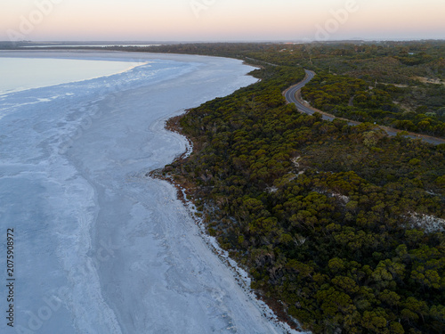 Aerial view of Pink Lake with salt crust shoreline, adjacent to native bushland and a winding coastal road under a soft twilight sky in Esperance, Western Australia, Australia.