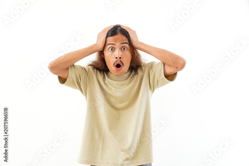 Canvastavla A portrait of a surprised young man with long hair, wearing a beige t-shirt, looking at the camera with his mouth open and hands on his head, isolated on a white background