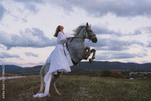 Fotografía Woman horse rearing majestic white steed against dramatic cloudy sky at sunset
