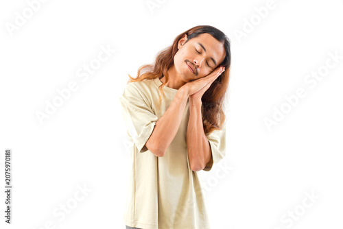 Slika na platnu A young man with long hair and a mustache poses with his hands pressed together against his cheek, mimicking a sleeping gesture, isolated on white