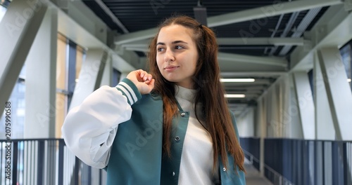 Caucasian woman on steel pedestrian bridge wearing teal varsity jacket and backpack, headphones around neck, thoughtful gaze with subtle smile, modern truss architecture and glass windows, candid