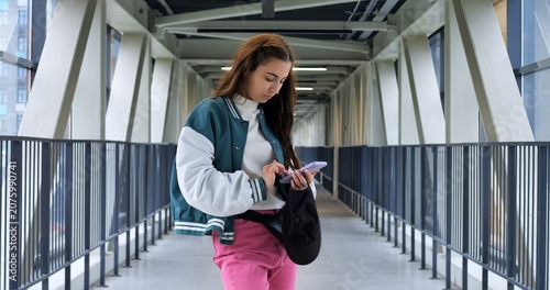 Young woman verifying eticket on smartphone while standing on pedestrian bridge, focused eyes scanning screen and holding small black bag, daylight through glass panels and modern transit context