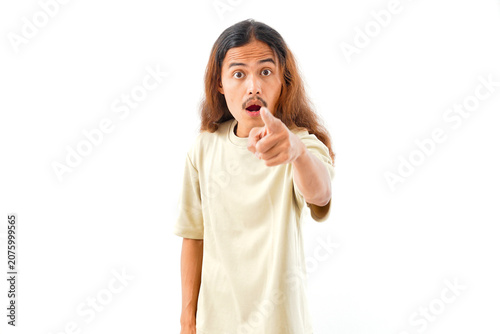 A young man with long hair and a mustache wearing a beige t-shirt looking surprised and pointing directly at the camera, isolated on a white background.