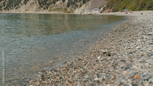Peaceful coastal scene with shallow clear water lapping against gray pebble shore. Background shows green cliff face distant beachgoers. Soft natural lighting, muted tones, serene mood