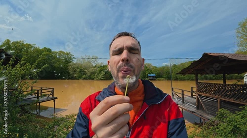 A man in a red jacket blows dandelion seeds and smiles outdoors near a muddy river. Wooden docks and green trees frame the scene under a bright blue sky with light clouds. Slow motion