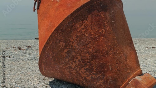 A weathered, rusted orange buoy rests on a gray pebbly seashore under a cloudy sky. The textured surface shows signs of decay. The soft natural light enhances the gloomy coastal atmosphere
