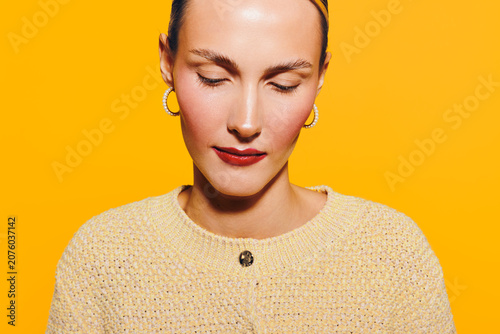 Fotografija Young woman with closed eyes and red lipstick wears yellow sweater and gold earrings on bright yellow background