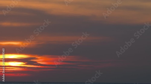 Distant airplane flying through dark clouds during dramatic red sunset over calm ocean, wide shot