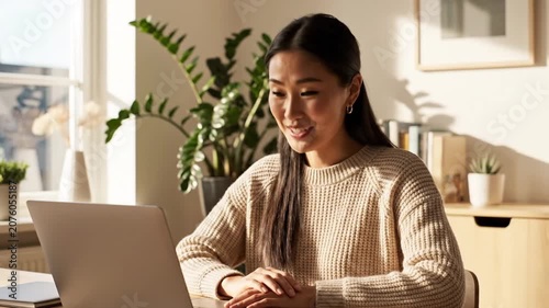 Smiling Asian woman uses laptop at sunny home office desk