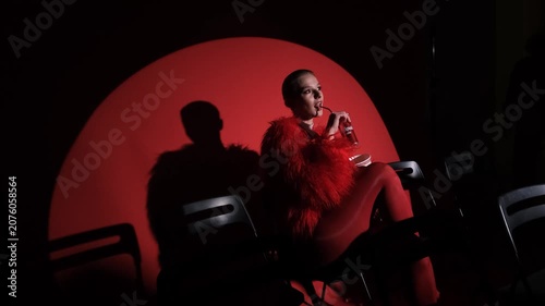Portrait of a beautiful young woman in a red fur coat against a red background, posing for a cinema-style photo. The woman smiles beautifully, sitting on chairs in the light of a projector during a
