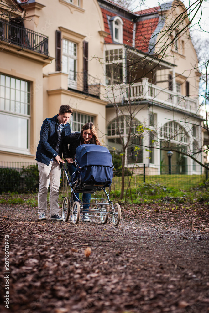 Obraz premium Junges Paar schiebt einen Kinderwagen auf einem Weg vor einer eleganten Altbauvilla, KI generiert