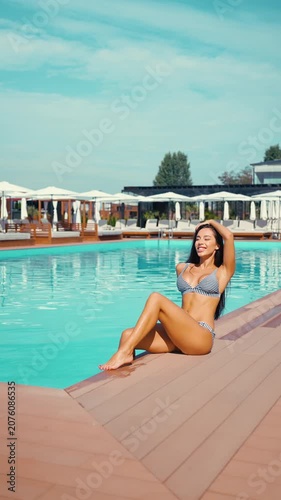 Beautiful young woman in striped bikini sitting by swimming pool. Smiling female enjoying summer vacation at luxury resort on sunny morning