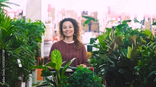 A happy woman with curly hair buys potted flowers at a garden center. The woman carries the flowers in a shopping cart.