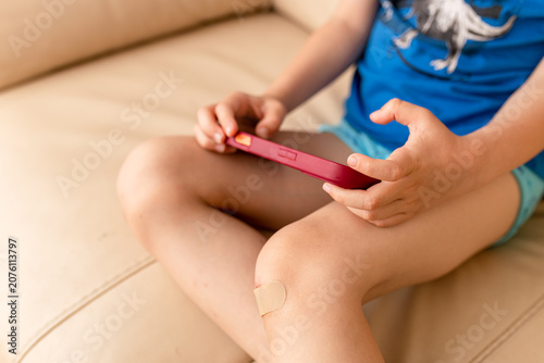 Child playing game on smartphone while sitting on couch with adhesive bandage on knee. Hurt kid recovering at home. Healthcare, injury, childhood, media addiction, leisure activity concept.