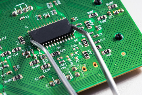 Tweezers holding a microchip processor on a green circuit board.