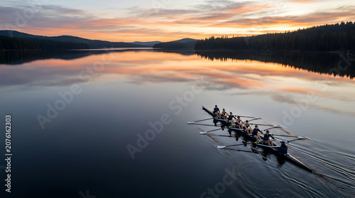 Aerial view of a rowing shell with a crew of eight rowers and a coxswain gliding across a calm, reflective lake during a vibrant sunrise surrounded by dense evergreen forest hills.
