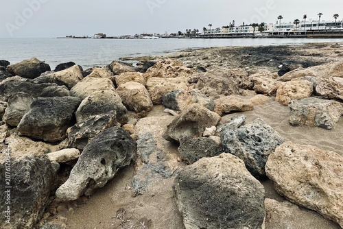 Beach in Kato Paphos, Cyprus with rocks and background promenade.
