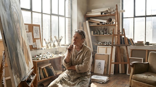 A thoughtful artist works quietly in a bright, naturally light-filled creative studio, surrounded by unfinished sketches, textured canvases, and a scattering of inspirational tools