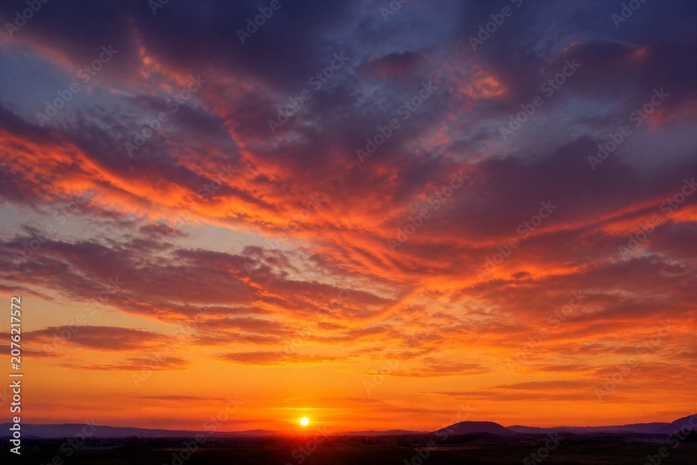Fototapeta premium Sun dips below distant hills, painting sky in fiery hues. Crimson clouds swirl with deep purples and golds. Horizon glows warmly as light fades into dusk