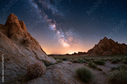 Dramatic night sky arches over rugged desert terrain. Milky Way glows with cosmic stars above jagged rock formations. Warm sunset hues linger near the horizon's edge. 