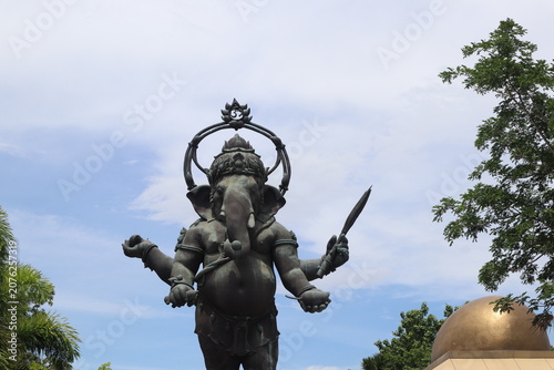 Fotografie Ganesha Statue Against Sky: A grand bronze statue of the Hindu deity Ganesha, with his characteristic elephant head and multiple arms, stands majestically against a partly cloudy blue sky