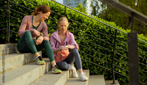 Fotografie Two women sit on steps in a park and check fitness watches during a workout brea