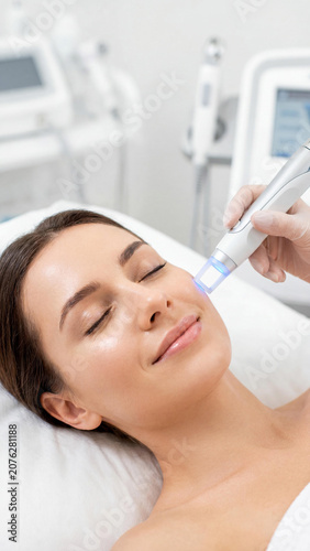 Smiling young Caucasian woman receiving a professional facial hydro-peeling skin treatment at a beauty clinic. Cosmetologist hand in glove holding a modern blue light laser device.