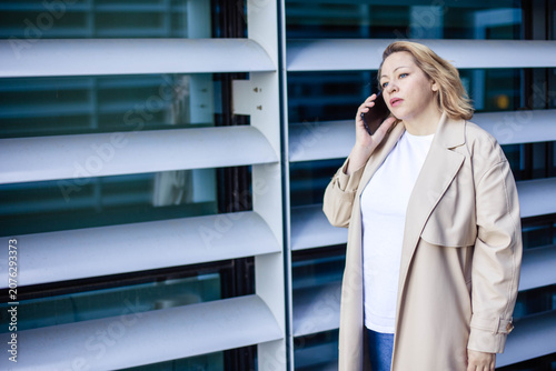 Plus-size woman in beige trench coat talking on smartphone while standing near modern building with horizontal blinds and glass windows in urban setting