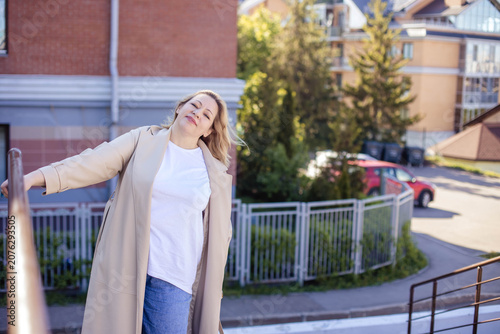 Caucasian woman in beige coat poses outdoors on a sunny day, standing on a staircase with residential buildings and greenery in the background