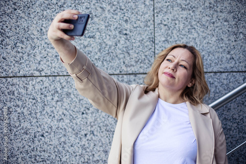 Caucasian woman taking a selfie with smartphone while standing against a textured stone wall, wearing a light-colored blazer and white shirt, smiling at the camera