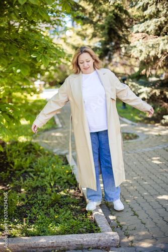 Woman in beige trench coat and white t-shirt stands on a pathway surrounded by greenery, enjoying a sunny day outdoors in a park setting