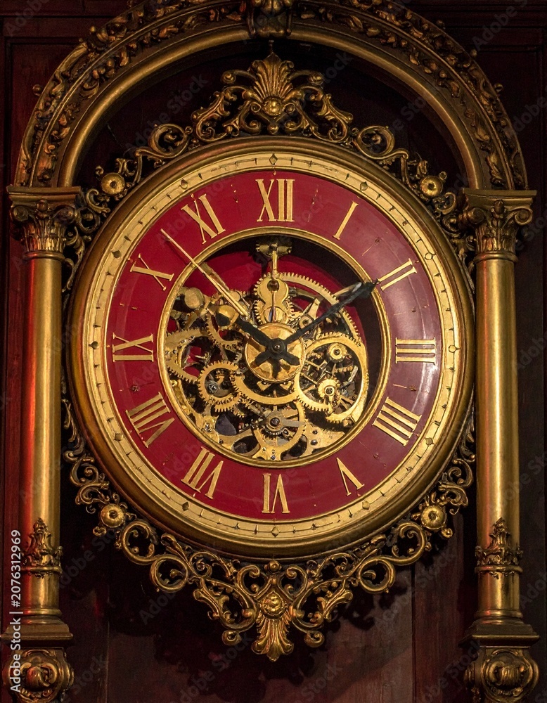 Obraz premium Magnificent baroque clock with exposed brass gears and Roman numerals, deep crimson enamel face, intricate gold filigree carvings, warm dramatic lighting.