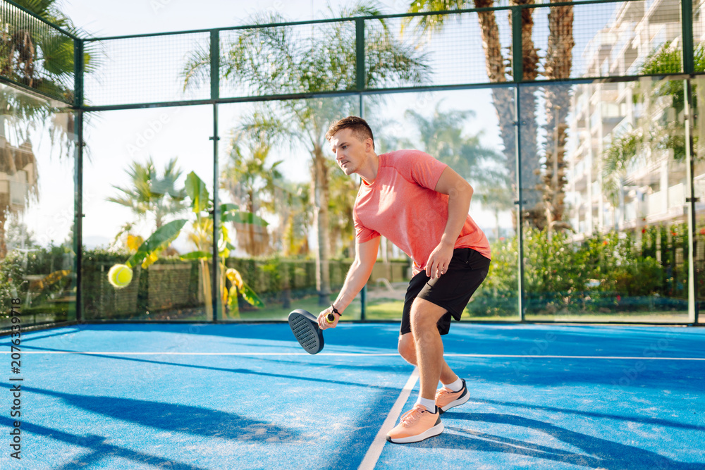 Obraz premium A young man on a blue tennis court holding a racket and ball. A man with a racket and ball practices on a paddle tennis court. Conceptual images of sports, activity, and weekends.