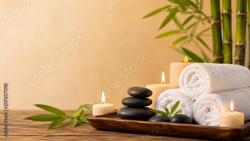 Stacked dark smooth stones and rolled white towels with lit candles and bamboo stalks on a wooden tray