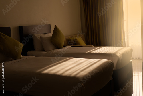 Cozy hotel room interior featuring twin beds with white linens, illuminated by warm golden sunlight streaming through the window blinds, creating a relaxing afternoon atmosphere