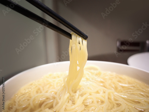 Close-up of a pair of black chopsticks lifting hot, yellow ramen noodles from a white ceramic bowl filled with flavorful broth