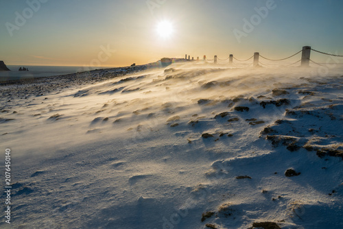 Strong gale force wind blowing spindrift snow over frozen ground during winter sunset, Dyrholaey, Iceland