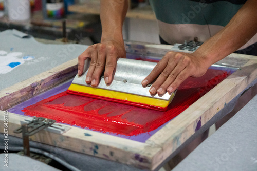 Photography Hand uses a squeegee with a yellow rubber blade to push red ink through a mesh screen