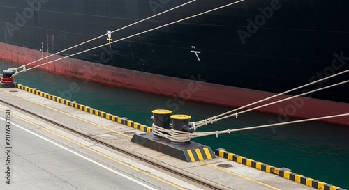 Large cargo ship secured to dock by thick ropes on mooring bollards at bustling port