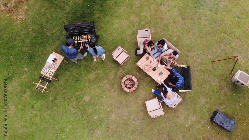 Fototapeta premium Aerial top-down view of a group of friends enjoying an outdoor barbecue party and camping on a green lawn, featuring a charcoal grill, wooden dining table, chairs, and a fire pit