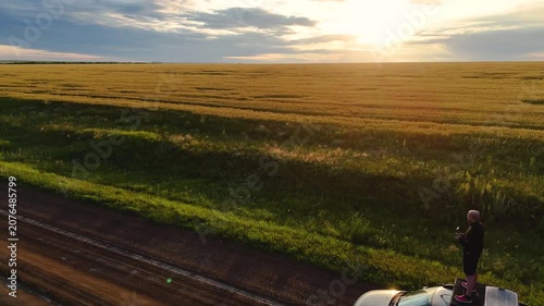A young man stands on the roof of a car in a wheat field during the rain at sunset