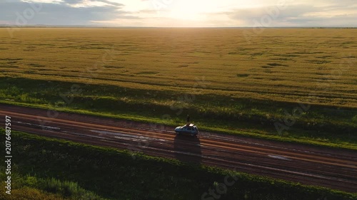 A young man stands on the roof of a car in a wheat field during the rain at sunset