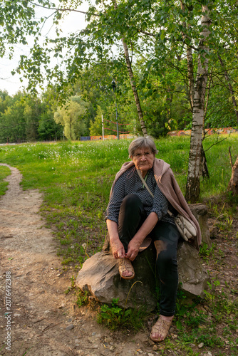 Senior woman sitting on a large stone while fastening her sandal during a peaceful walk through a birch woodland. Active aging, independent lifestyle, outdoor recreation, mobility, healthy retirement,