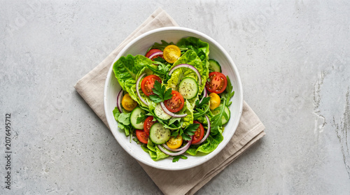 Photos Top down view of a fresh garden salad with lettuce, tomatoes, cucumbers, and onions in a white bowl, placed on a grey concrete background