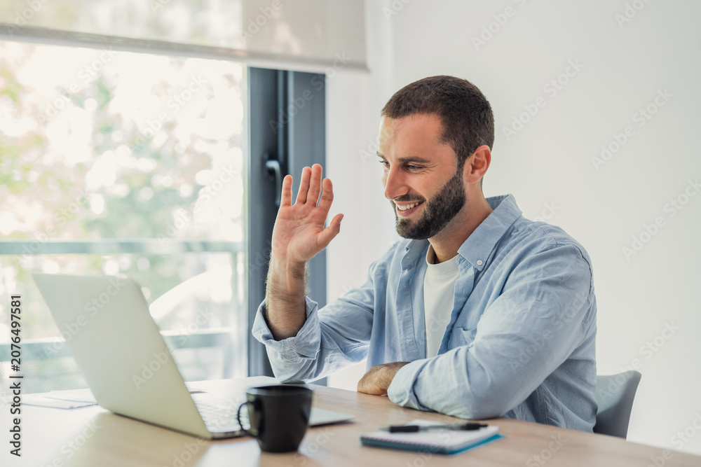 Fototapeta premium Young man smiling and waving to a laptop screen during a video call, communicating remotely from his modern home office setup while engaging in virtual conversations
