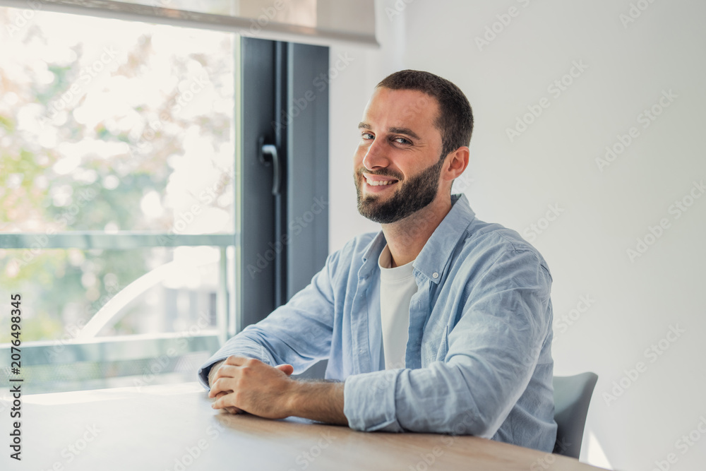 Fototapeta premium Smiling young man in a light blue shirt sits at a home office desk near a sunny window, conveying relaxed confidence, remote work lifestyle, productivity and approachable professionalism.