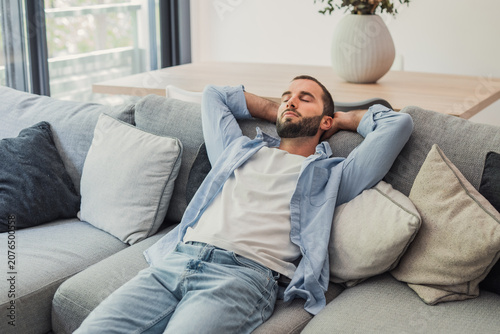 Fotografía Young man wearing a casual shirt and jeans resting on a comfortable gray sofa, n