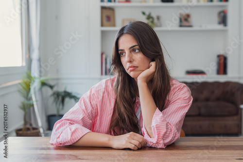 Tableau sur toile Young woman sitting at a modern wooden table, resting her chin on her hand, look